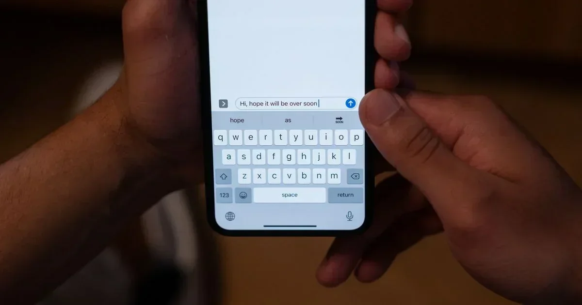Hands holding a smartphone displaying a messaging app with the text "Hi, hope it will be over soon" typed into the input field above a QWERTY keyboard.